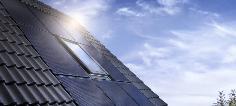 Solar roof tiles installed on a house slope under a bright blue sky with clouds, showing a small skylight window.
